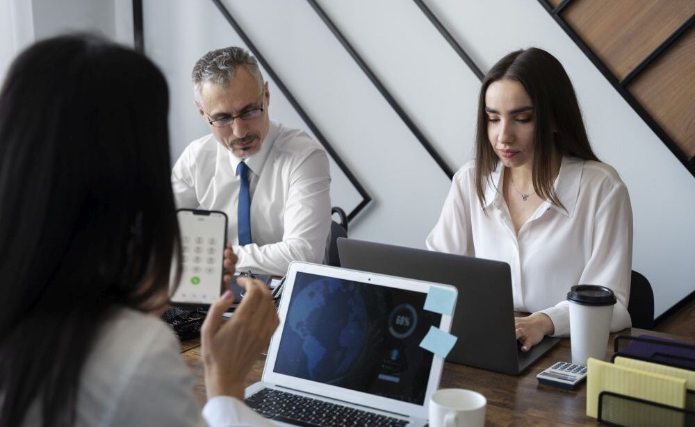 Three people are sitting at a table with laptops, a phone, coffee cups, and office supplies, collaborating for a tech search agency in a modern office setting.