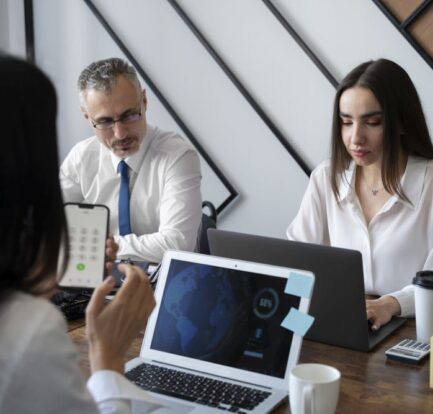 Three people are sitting at a table with laptops, a phone, coffee cups, and office supplies, collaborating for a tech search agency in a modern office setting.