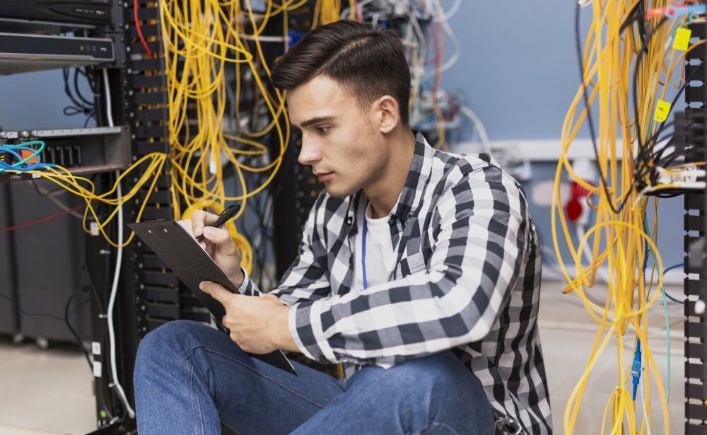 A network cabling contractor in a checkered shirt sits on the floor of a server room, writing on a clipboard, surrounded by an array of network cables and server racks.