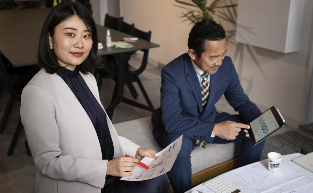 Two professionals in business attire work together; one woman holds documents and a pen, while a man checks data on a tablet, discussing PSG-claimable IT and security packages for first-time grant applicants in Singapore at a table with papers and coffee.