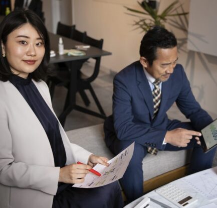 Two professionals in business attire work together; one woman holds documents and a pen, while a man checks data on a tablet, discussing PSG-claimable IT and security packages for first-time grant applicants in Singapore at a table with papers and coffee.