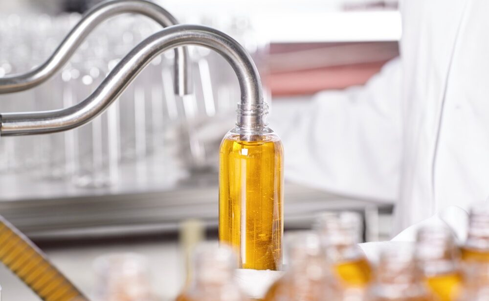 A machine dispenses a liquid, possibly oil, into a clear plastic bottle on a production line. A person in a lab coat is present in the background.