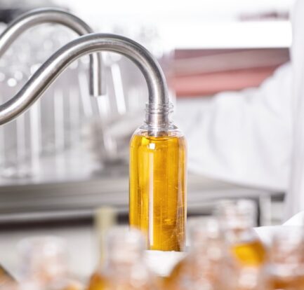 A machine dispenses a liquid, possibly oil, into a clear plastic bottle on a production line. A person in a lab coat is present in the background.